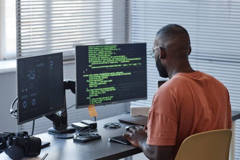 Professional black man sitting in front of two computer screens with code